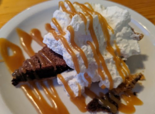A slice of chocolate dessert topped with whipped cream and drizzled with caramel sauce on a white plate.