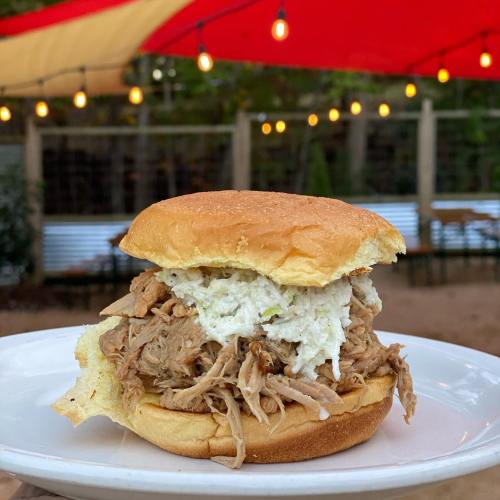 A pulled pork sandwich with coleslaw on a soft bun, served on a plate outdoors with string lights in the background.