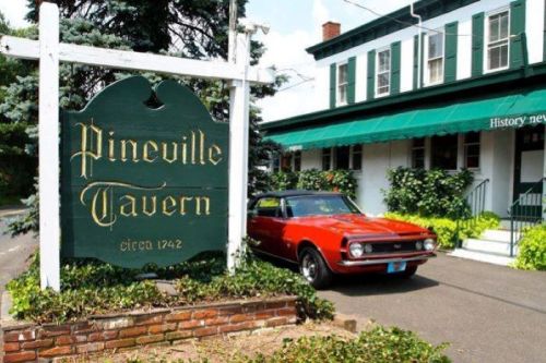 A red vintage car parked in front of Pineville Tavern, a historic building with a green sign and lush greenery.
