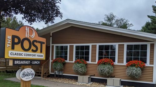 A cozy restaurant exterior with a sign reading "The Post" and flower planters by the windows, promoting breakfast and lunch.
