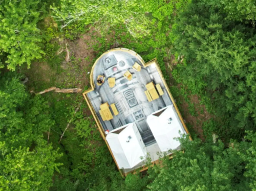 Aerial view of a unique cabin nestled in a forest, featuring a circular design and wooden deck with seating.