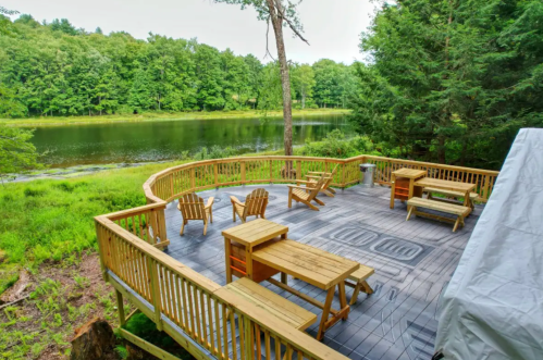 A wooden deck with chairs and tables overlooks a serene pond surrounded by lush greenery.