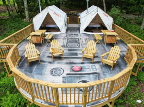 Two white tents on a wooden deck with Adirondack chairs and tables, surrounded by greenery.