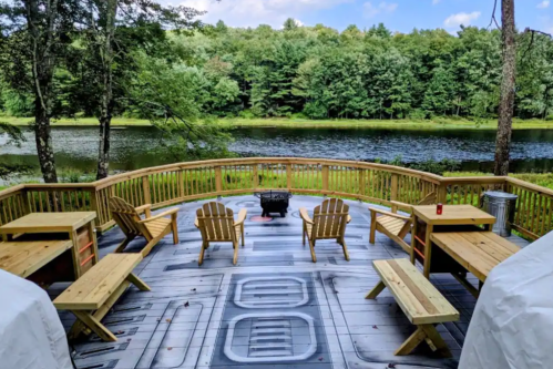 A scenic deck with wooden chairs and tables overlooking a calm lake surrounded by trees under a blue sky.