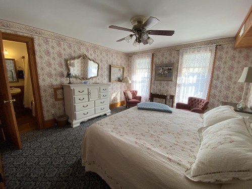 Cozy bedroom with floral wallpaper, a large bed, vintage furniture, and two armchairs by the window.