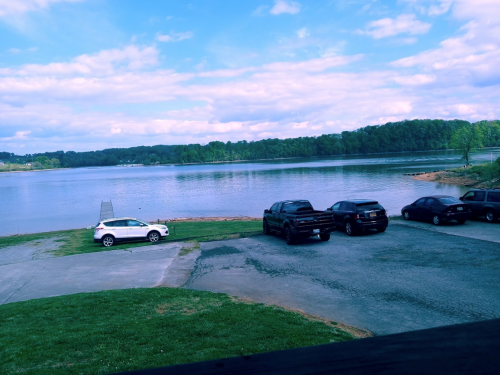 A serene lake view with several parked cars along the shore and a cloudy blue sky above.