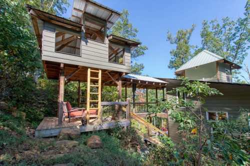 A unique two-story house nestled among trees, featuring a deck and a ladder leading to an upper level.