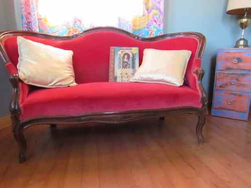 A vintage red sofa with two pillows, next to a colorful artwork and a small wooden nightstand.