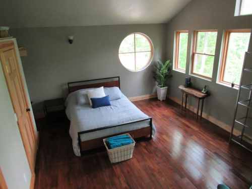 A cozy bedroom with a round window, wooden floors, a bed with blue accents, and a small table by the window.