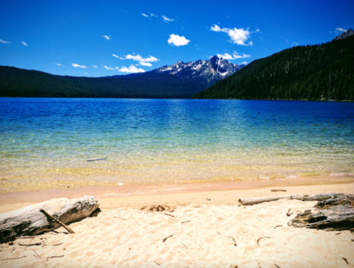 A serene lake with clear blue water, sandy shore, and mountains in the background under a bright blue sky.