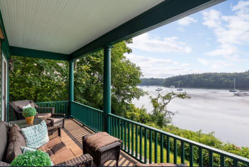 A cozy porch with wicker chairs overlooks a calm river and boats, surrounded by lush greenery and a clear sky.