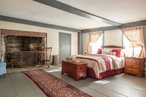 Cozy bedroom with a wooden bed, rocking chair, and a stone fireplace, featuring warm lighting and a patterned rug.