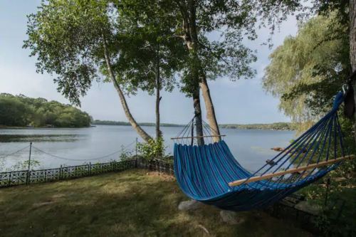 A colorful hammock hangs between trees by a calm lake, surrounded by lush greenery and a peaceful atmosphere.