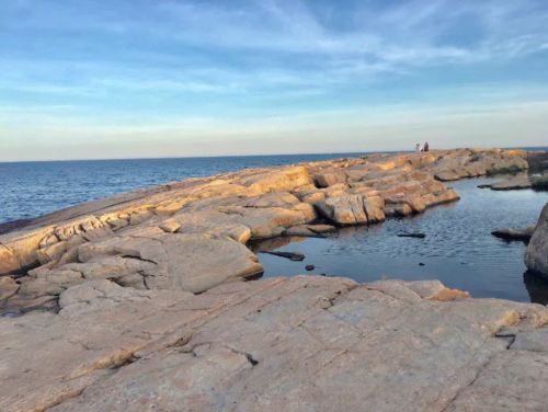 Rocky shoreline with a calm sea and a small pool of water, under a clear blue sky. Two people are visible in the distance.