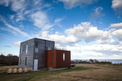 Modern two-story house with a mix of wood and metal exterior, set against a cloudy sky and grassy landscape.