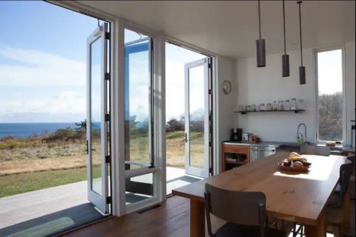 Bright kitchen with open doors leading to a scenic view of the ocean and grassy landscape. Modern design and wooden table.