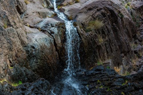 A small waterfall cascading over rocky terrain, surrounded by rugged cliffs and sparse vegetation.