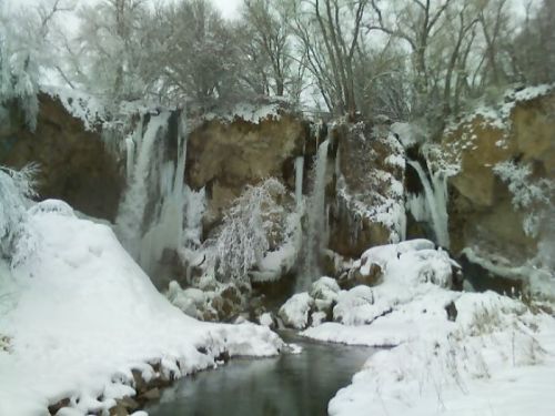 A snowy landscape featuring frozen waterfalls surrounded by trees and a calm river.