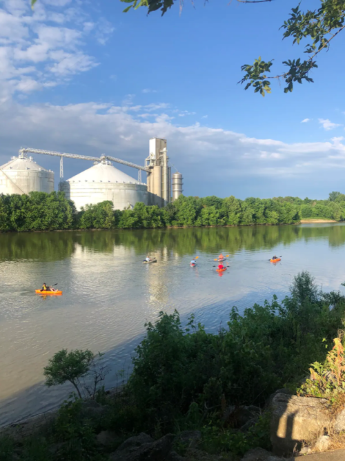 Kayakers paddle on a river near silos and lush greenery under a partly cloudy sky.