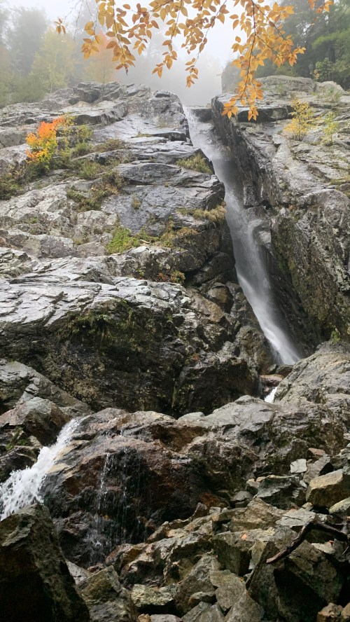 A misty waterfall cascading down rocky cliffs, surrounded by autumn foliage and rugged terrain.