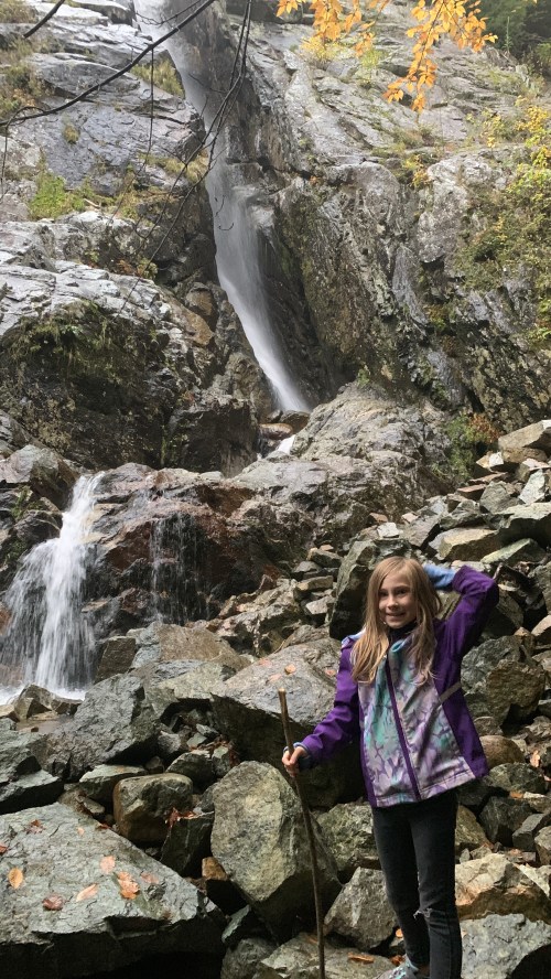 A girl stands on rocky terrain near a waterfall, holding a stick, surrounded by autumn foliage.