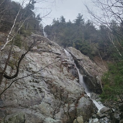 A cascading waterfall flows down rocky terrain, surrounded by trees and a misty atmosphere.