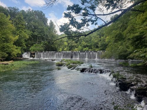 A serene river scene featuring a waterfall surrounded by lush green trees and a clear blue sky.