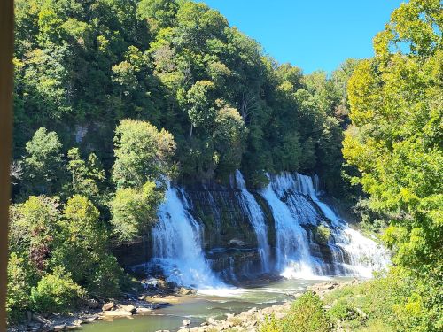 A scenic view of a waterfall cascading down rocky cliffs, surrounded by lush green trees under a clear blue sky.