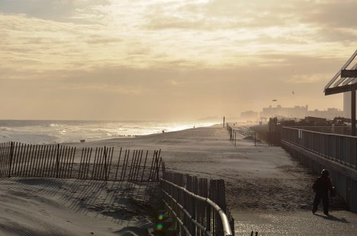 A serene beach scene at sunset, with gentle waves, distant buildings, and a person walking along the shore.