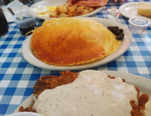 A plate of fried chicken with gravy, a large pancake, and sides of potatoes and ham on a checkered tablecloth.