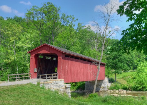 A red covered bridge spans a calm stream, surrounded by lush green trees and a clear blue sky.