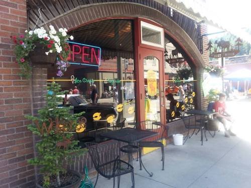 A cozy café with an "OPEN" sign, outdoor seating, and colorful flower pots, bustling with people on a sunny day.