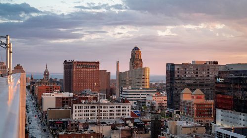 A city skyline at sunset, featuring various buildings and a cloudy sky over a bustling urban area.