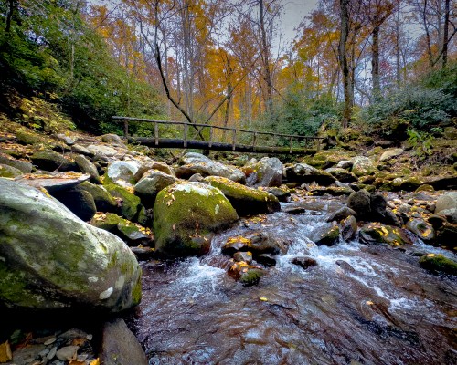 A serene stream flows over rocks, with autumn foliage and a wooden bridge in the background.