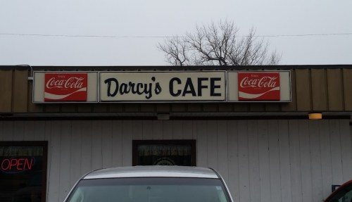 Sign for Darcy's Cafe featuring Coca-Cola logos, with a gray car parked in front and a cloudy sky above.
