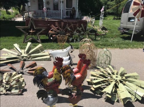 A collection of decorative metal roosters and windmill parts displayed outdoors, with a house in the background.