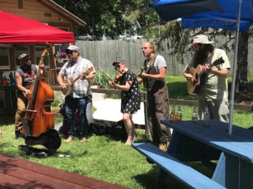 A group of five musicians play instruments outdoors, surrounded by greenery and colorful decorations.