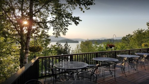 A scenic view from a deck overlooking a lake, surrounded by trees and mountains, with tables set for dining.