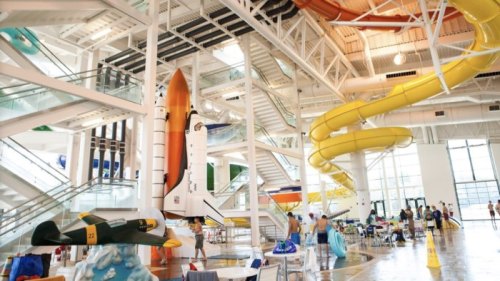 Indoor water park featuring a space shuttle, rocket slide, and colorful water slides with people enjoying the space.