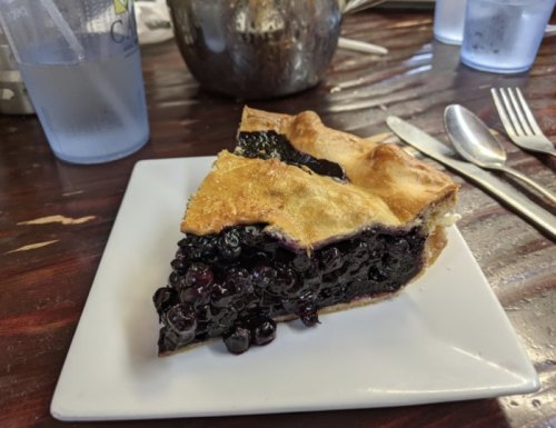 A slice of blueberry pie on a white plate, with a golden crust and a glossy filling, set on a wooden table.