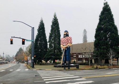 A large statue of a lumberjack stands near a street intersection, surrounded by tall trees and a foggy sky.