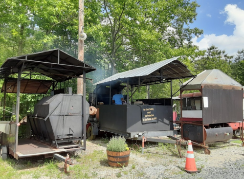 Two barbecue smokers with smoke rising, surrounded by greenery and a rustic setting. A person is visible near the smokers.