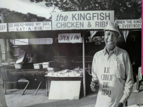 A smiling chef in an apron stands outside a barbecue restaurant, "The Kingfish Chicken & Ribs," with signs and food stalls.
