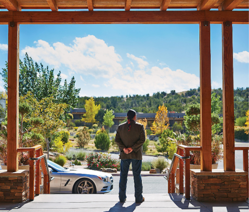 A person stands with hands clasped, looking out at a scenic view of trees and a car parked nearby.