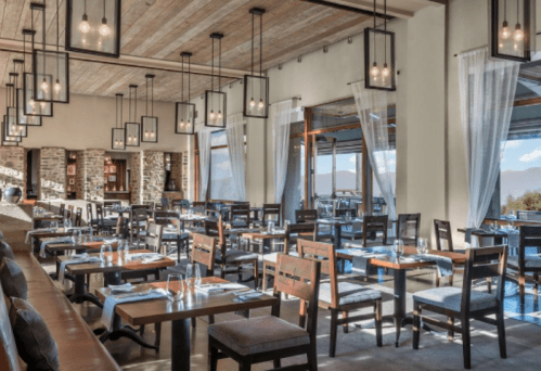 Modern restaurant interior with wooden tables, chairs, and large windows, featuring pendant lighting and a cozy atmosphere.