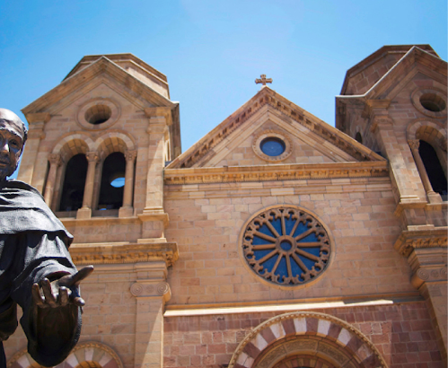 A statue in front of a historic stone church with large windows and a cross on top, under a clear blue sky.