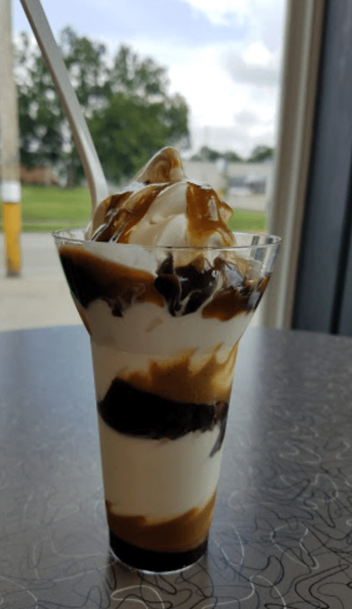 A clear cup filled with layers of vanilla ice cream, chocolate sauce, and caramel, topped with whipped cream.
