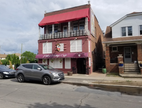 A two-story café with a red awning and balcony, surrounded by parked cars and residential buildings.