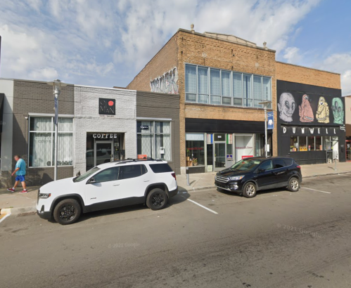 A street view featuring two buildings with shops, parked cars, and pedestrians on a sunny day.