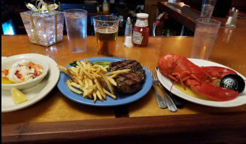 A table with a lobster, steak with fries, and a dessert, alongside drinks and condiments in a restaurant setting.